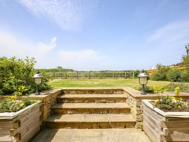 A garden with stone steps leading to a lawn with wooden planters and fence at Fairview Cottage in Milton-Under-Wychwood