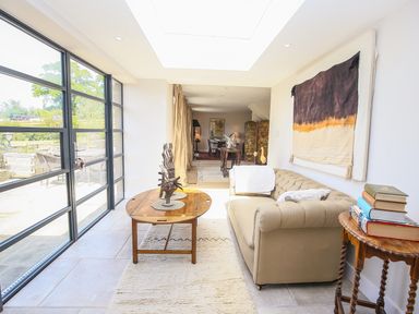 A sitting area with a beige sofa a wooden coffee table with abstract sculptures and a side table with books next to large windows at Fairview Cottage in Milton-Under-Wychwood