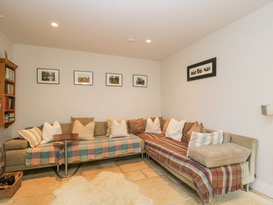 A living room with a sectional sofa covered in blankets and cushions and framed pictures on the wall at The Lodge in Coln St Aldwyns