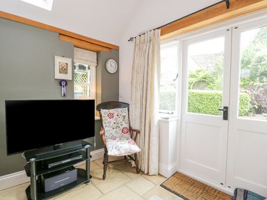 A corner of a living room with a TV on a glass stand a wooden chair with floral cushions and double doors leading outside at The Lodge in Coln St Aldwyns