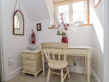 A desk with a chair and a vase of flowers next to a small drawer and mirror on the wall at The Lodge in Coln St Aldwyns