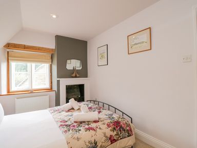 A bedroom with a bed with floral bedding towels on the bed a window a radiator paintings on the wall and a fireplace at The Lodge in Coln St Aldwyns