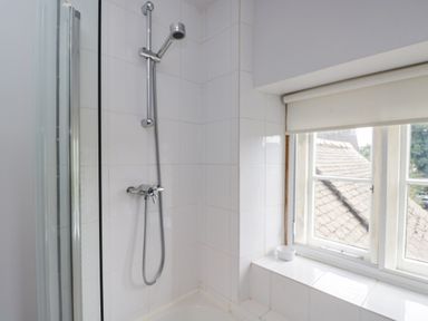 A shower area with a showerhead and tiled walls next to a window with a roller blind at The Lodge in Coln St Aldwyns