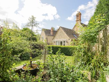 A garden with green plants and trees in front of stone houses at The Lodge in Coln St Aldwyns