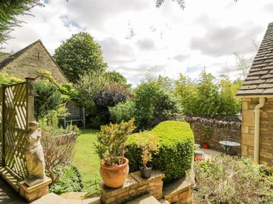 A garden with a stone statue and potted plants near a wooden gate and stone wall with outdoor seating at The Lodge in Coln St Aldwyns