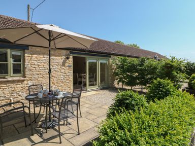An outdoor patio area with a metal table and chairs under a large umbrella in front of a stone building with green windows and doors at Ewecub Barn in Burford