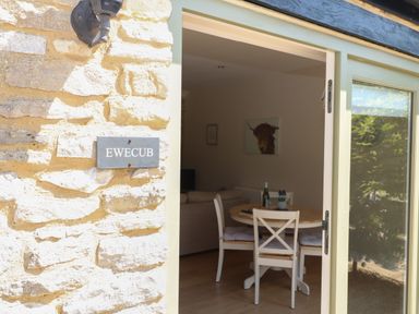 A dining area with a round wooden table and white chairs inside a stone building at Ewecub Barn Burford