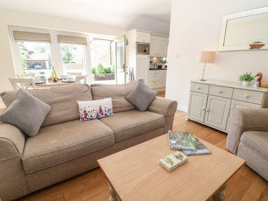 A living room with a beige sofa, a wooden coffee table with magazines, a sideboard with a lamp and plants at Ewecub Barn in Burford