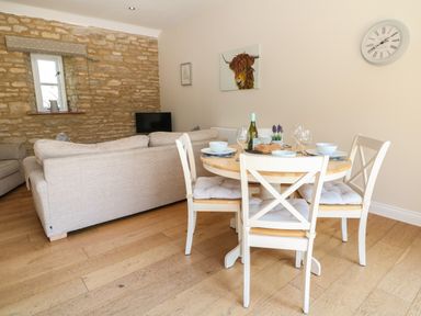 A dining area with a round wooden table and four white chairs next to a living room with beige sofas and a stone wall at Ewecub Barn Burford