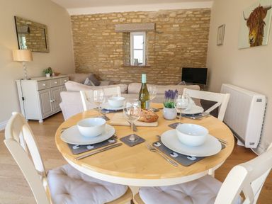 A dining area with a round wooden table set with bowls wine glasses cutlery and a loaf of bread with a sitting area and stone wall in the background at Ewecub Barn in Burford
