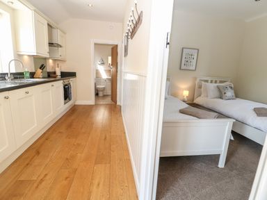 A kitchen with wooden floor and white cabinets next to a room with two single beds and a lamp at Ewecub Barn in Burford