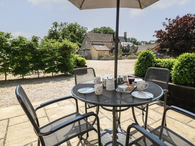 An outdoor patio table with cups plates and a scone under an umbrella with stone buildings and greenery in the background at Ewecub Barn in Burford