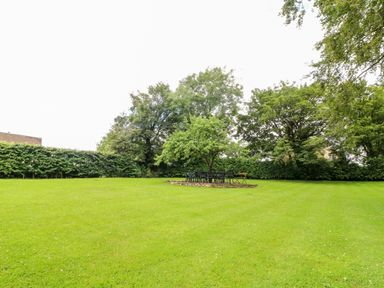 A large grassy garden with trees and a circular paved seating area with metal chairs and a bench at South Hill Farmhouse in Stow-On-The-Wold