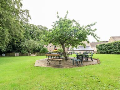 An outdoor patio area with tables and chairs under a tree in a grassy garden with buildings in the background at South Hill Farmhouse in Stow-On-The-Wold
