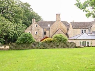 A stone house with multiple chimneys behind a wooden fence and a green lawn at South Hill Farmhouse in Stow-On-The-Wold
