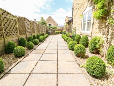 A stone paved pathway lined with round shrubs next to a stone house and a wooden fence at South Hill Farmhouse in Stow-On-The-Wold