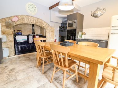 A kitchen with wooden dining table and chairs a black stove under a stone arch clock on wall and modern cabinets at South Hill Farmhouse in Stow On The Wold