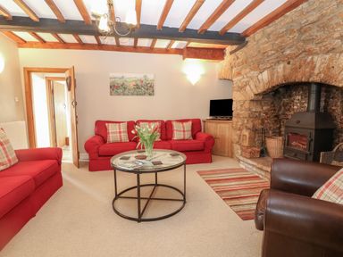 A living room with red sofas a round glass coffee table wooden beams and a stone fireplace at South Hill Farmhouse in Stow-On-The-Wold