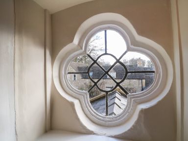 An exterior view of stone buildings through a four-lobed shaped window at South Hill Farmhouse in Stow-On-The-Wold