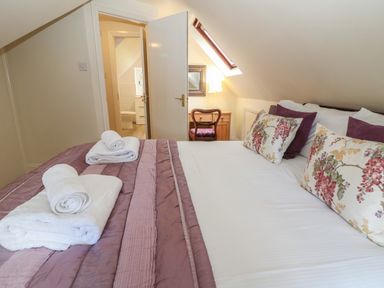 A bedroom with a bed covered with pillows and towels with a doorway showing a bathroom and a wooden desk with a chair and lamp at South Hill Farmhouse in Stow-On-The-Wold