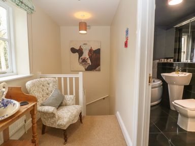 A landing area with a patterned armchair and wooden table near a window with a painting of a cow on the wall and a view into a bathroom with a sink toilet and bathtub at South Hill Farmhouse in Stow-On-The-Wold