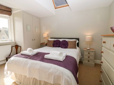 A bedroom with a double bed featuring white bedding purple throw and towels bedside tables with lamps a wardrobe a chair and a chest of drawers at South Hill Farmhouse in Stow-On-The-Wold