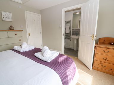 A bedroom with a bed covered with white and purple linens two rolled white towels on the bed a green dresser with a vase a wooden chest of drawers and an open door leading to a bathroom at South Hill Farmhouse in Stow-On-The-Wold