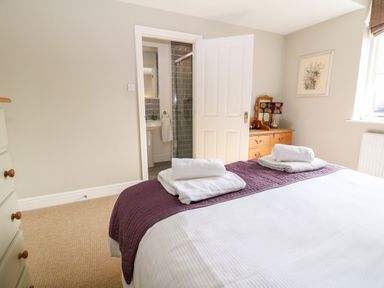 A bedroom with a bed with white and purple linens towels on the bed a wooden chest of drawers with a lamp and a picture on the wall at South Hill Farmhouse in Stow-On-The-Wold