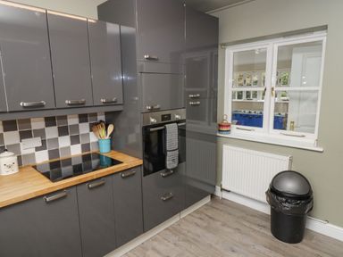 A kitchen with grey cabinets a built-in oven an electric stovetop wooden countertop and a window with a radiator below at South Hill Farmhouse in Stow-On-The-Wold