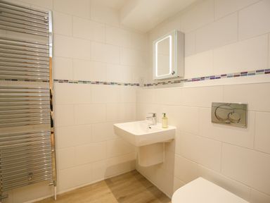 A bathroom with a wall-mounted sink with a soap dispenser a lit mirror above a toilet and a chrome towel radiator at 5 Burford Mews in Burford