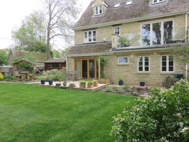A stone house with a tiled roof a wooden bench pots with plants on a patio and a green lawn with bushes at 5 Burford Mews in Burford