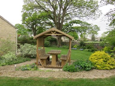 A wooden garden seating area with benches and a table surrounded by grass and plants at 5 Burford Mews in Burford