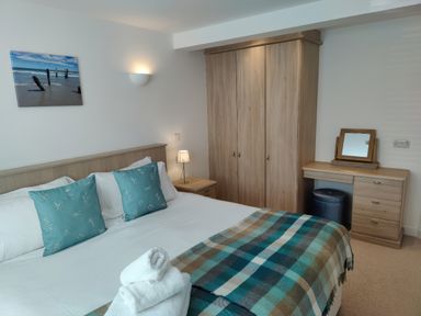 A bedroom with a bed with blue pillows and a plaid blanket a wooden wardrobe a dressing table with a mirror and a small lamp next to the bed at 5 Burford Mews in Burford