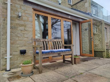 A wooden bench with a cushion on a stone patio outside a house with large glass doors at 5 Burford Mews in Burford