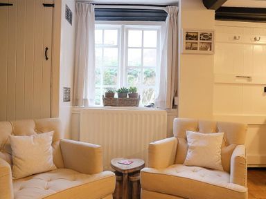 A sitting area with two beige armchairs a small wooden table between them and a window with potted plants at One Masons Court in Stratford upon Avon