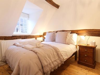 A bedroom with a double bed with beige bedding and pillows next to a wooden nightstand with a lamp at One Masons Court in Stratford upon Avon