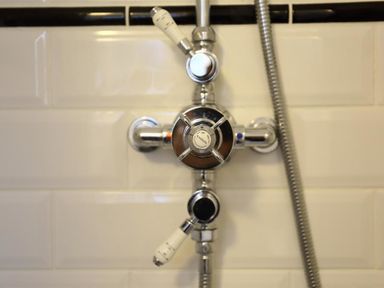 A close up of a shower control valve with white handles and a flexible metal hose attached on a tiled wall at One Masons Court in Stratford upon Avon