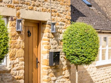 A wooden front door with black handle and knocker on a stone wall with two lights and a black mailbox beside a round trimmed bush at Orchard House in Stow-On-The-Wold