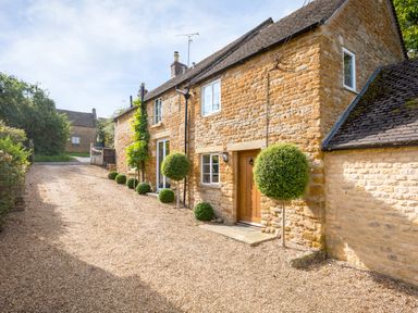 A stone exterior house with a wooden door and multiple spherical topiary plants along a gravel driveway at Orchard House in Stow-On-The-Wold