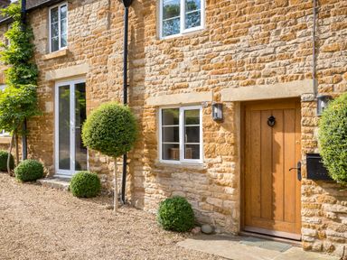 Exterior of a stone house with a wooden door and white framed windows with spherical and bushy plants at Orchard House in Stow-On-The-Wold