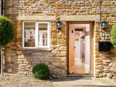 A stone exterior wall with a wooden door open to a hallway and two trimmed round bushes at Orchard House in Stow-On-The-Wold
