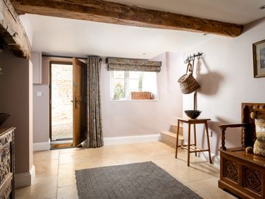 An entryway with a wooden door open to the outside a window with a basket on the sill a coat rack with a hanging basket a wooden table with a bowl a wooden bench with cushions and a patterned rug on the floor at Orchard House in Stow-On-The-Wold