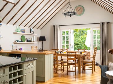A dining area with wooden chairs and table next to open French doors showing greenery outside at Orchard House in Stow-On-The-Wold