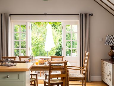 A dining area with wooden chairs and table set with breakfast items near open glass doors showing a garden at Orchard House in Stow-On-The-Wold