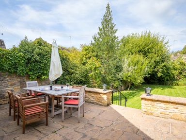 An outdoor patio with a table and six chairs with red cushions and a closed umbrella overlooking a garden with trees and grass at Orchard House in Stow On The Wold