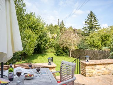 A patio with a wooden table set with wine glasses bottles and fruit bowl overlooking a garden with grass and trees at Orchard House in Stow On The Wold