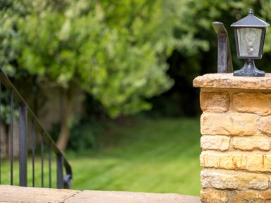 A stone wall with a black lantern on top and a metal handrail on the left with green grass and trees in the background at Orchard House in Stow-On-The-Wold