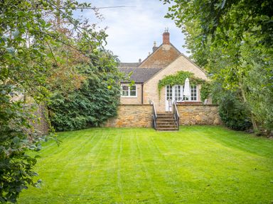 A stone house with a patio and stairs leading to a green lawn surrounded by trees and shrubs at Orchard House in Stow-On-The-Wold