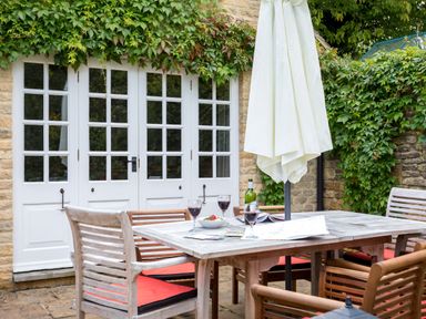 An outdoor patio area with a wooden table and chairs with red cushions white umbrellas and green ivy on the stone wall and white doors at Orchard House in Stow-On-The-Wold
