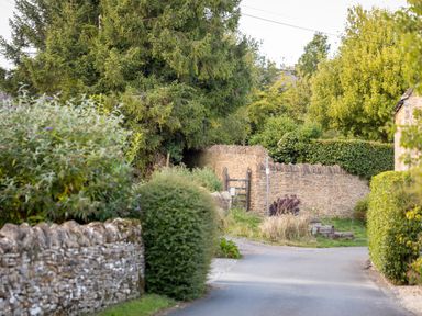 A country lane with stone walls, shrubs, trees, and a wooden gate at Orchard House in Stow-On-The-Wold